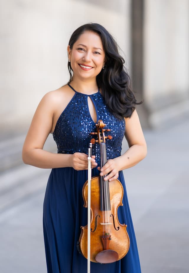 Sandra Morales Corrales holding a violin with a blue dress and behind is a rural background