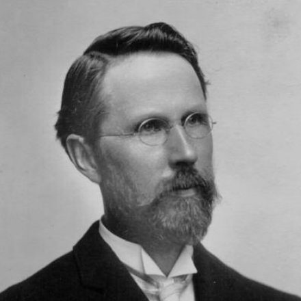 Silas Mason poses for the camera wearing round, thin-framed glasses, a white dress shirt and a black coat.