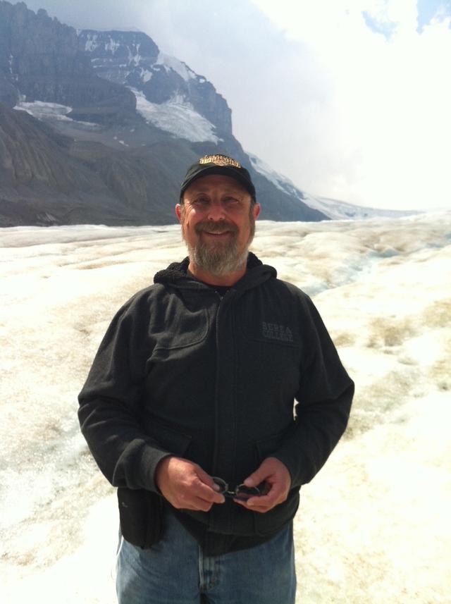 Ron Rosen portrait, wearing a black top, holding glasses, with a background of glaciers and mountains.