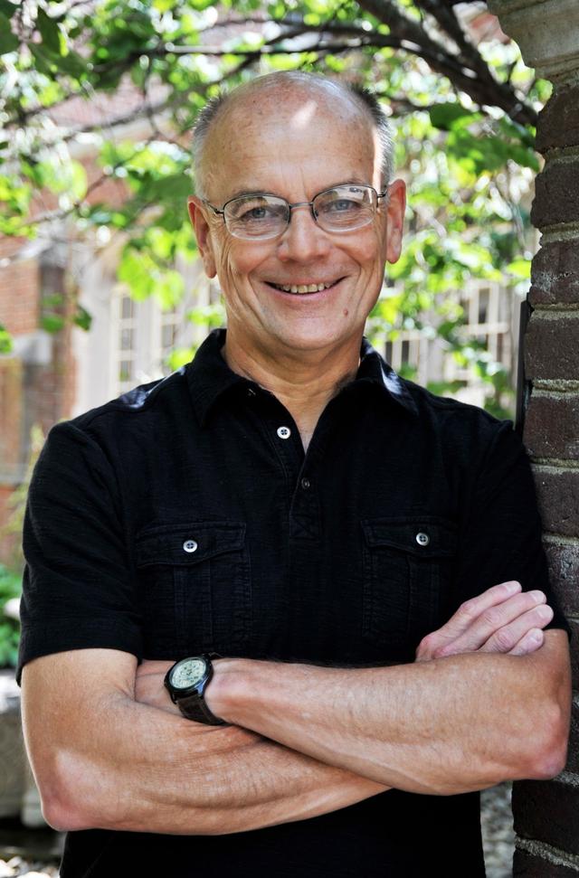 Steve Gowler headshot wearing a black shirt learning near a wall and in the background are trees full of greenery.