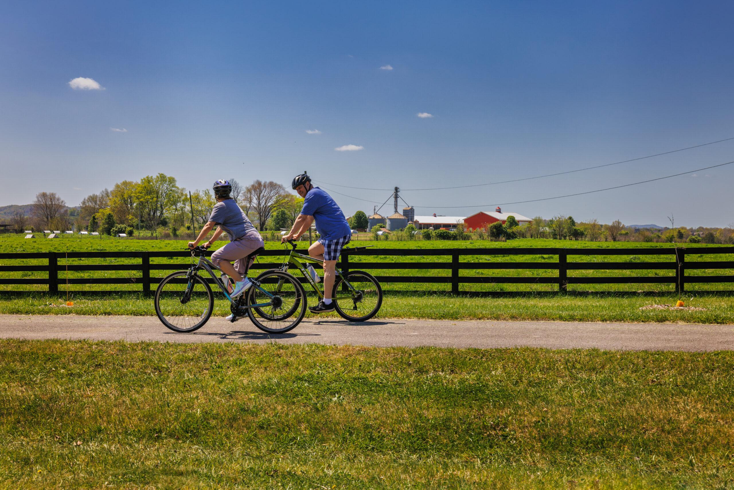 A picture of two people biking and there is a farm on the background and the sun is shining