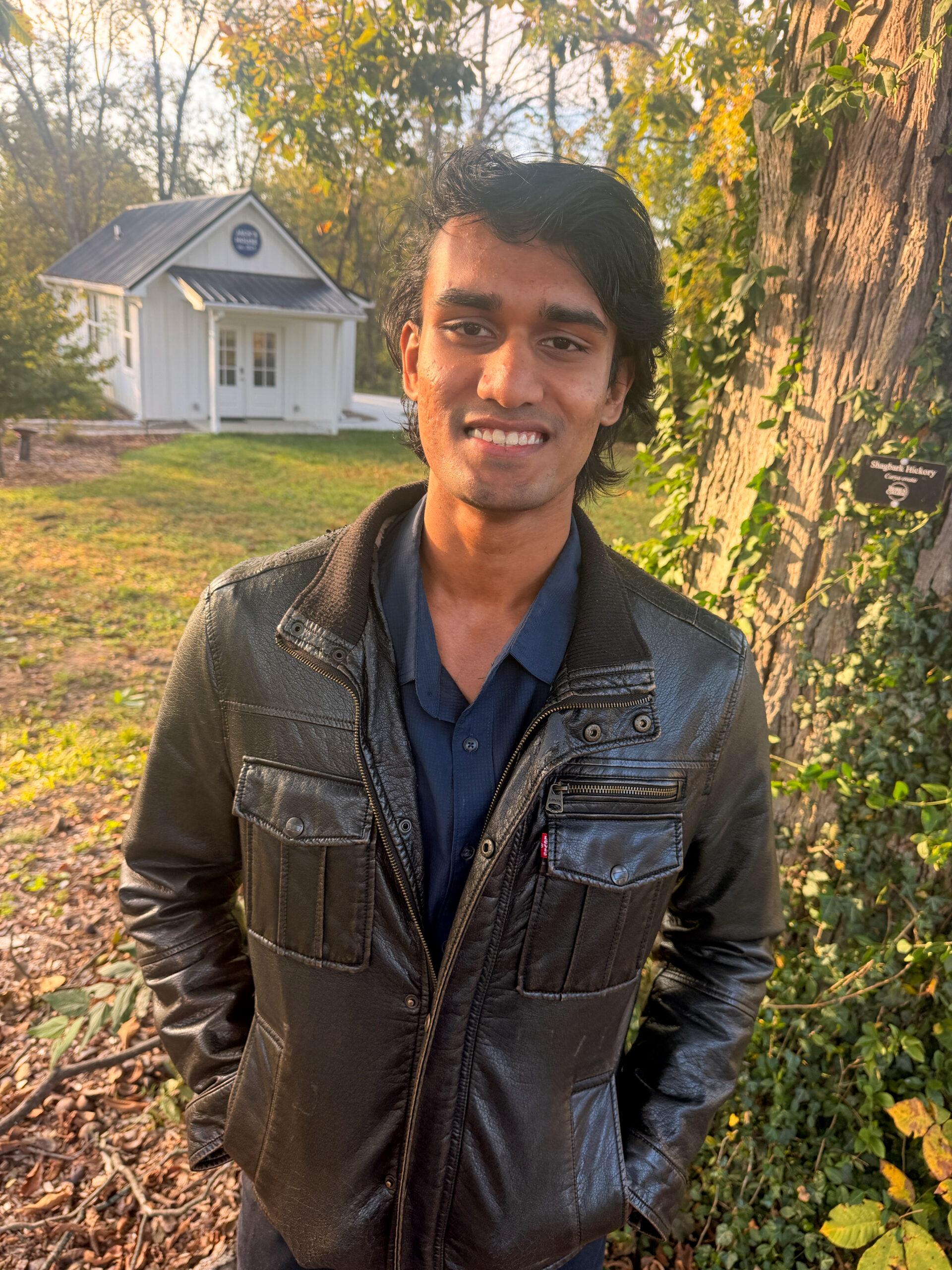 Person standing outdoors beside a large tree, wearing a black leather jacket and navy shirt, with a small white building (Jack's House) and autumn foliage in the background.