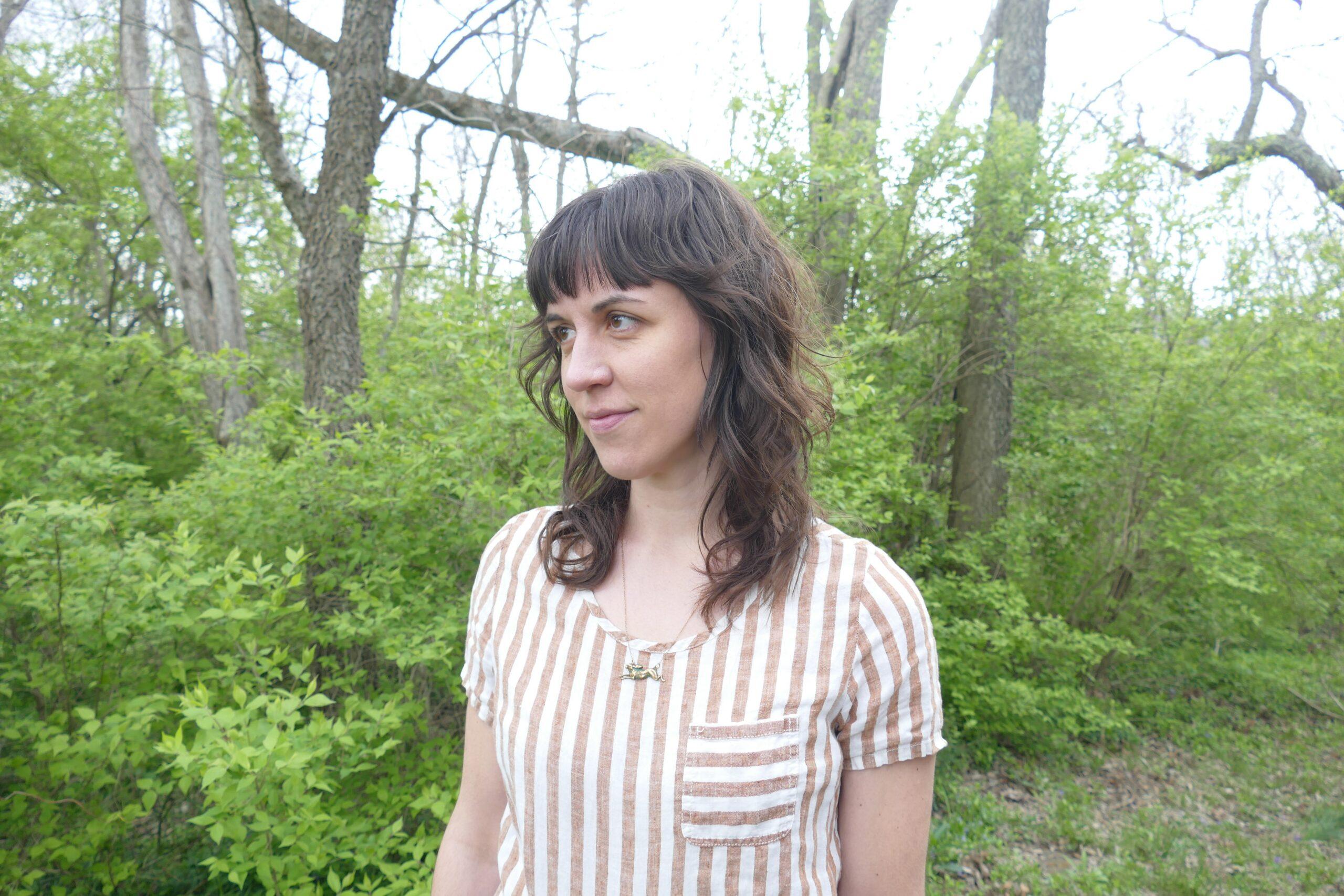 A woman with long, wavy brown hair standing in a forest setting.