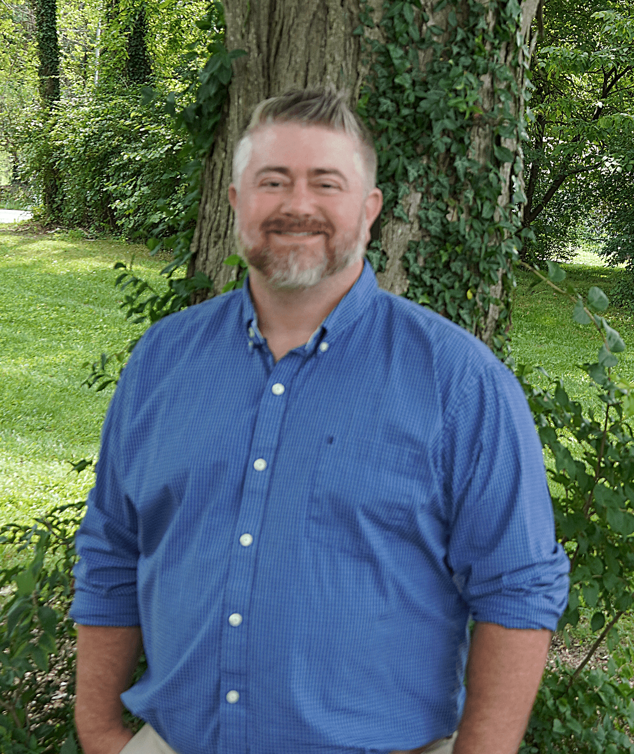 Brandon standing outdoors in front of a tree covered with ivy, wearing a blue button-down shirt.