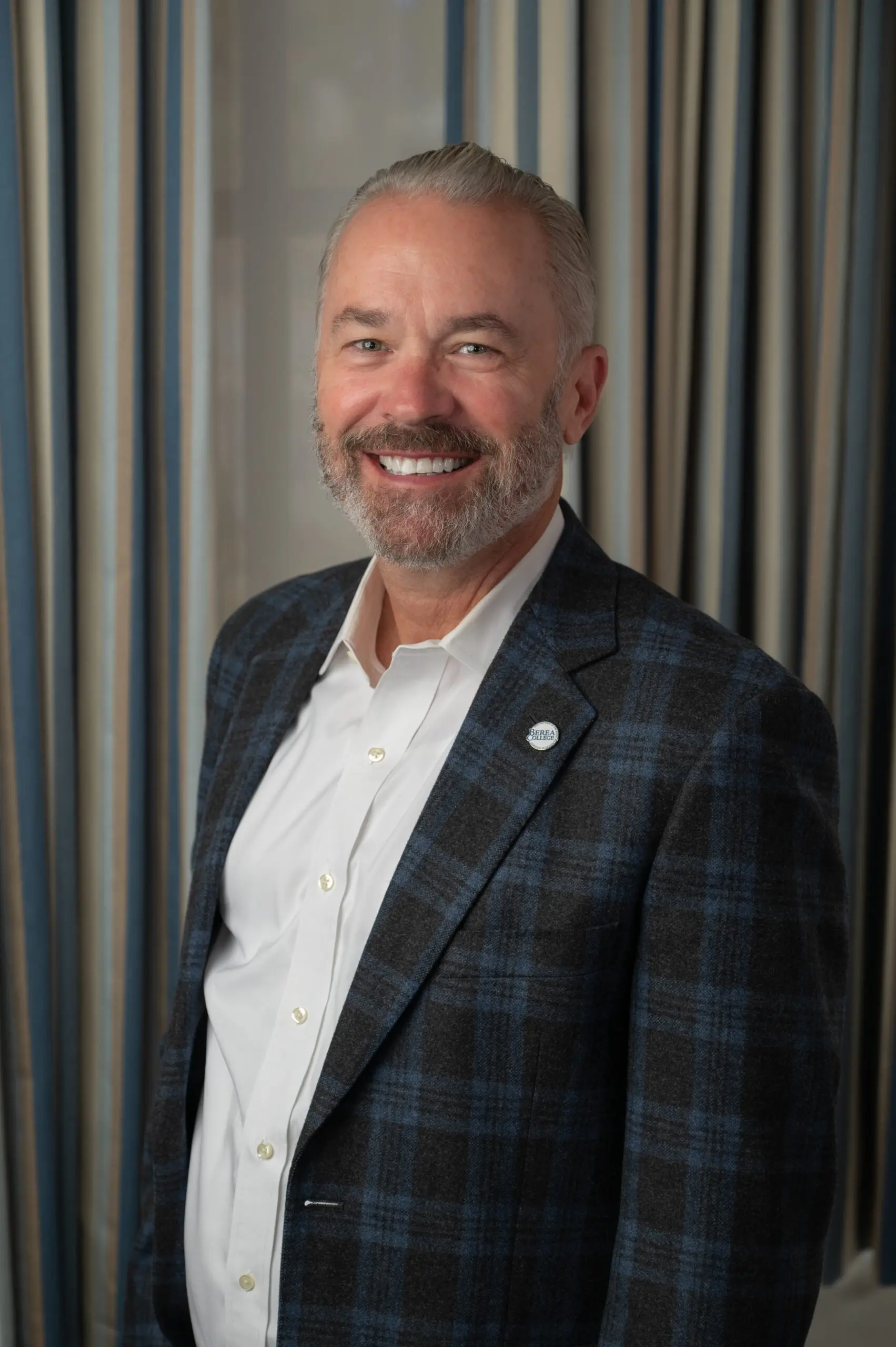 Chad Berry is standing in front of gray backdrop. He is smiling and wearing a blue and green checkered jacket with a white shirt.