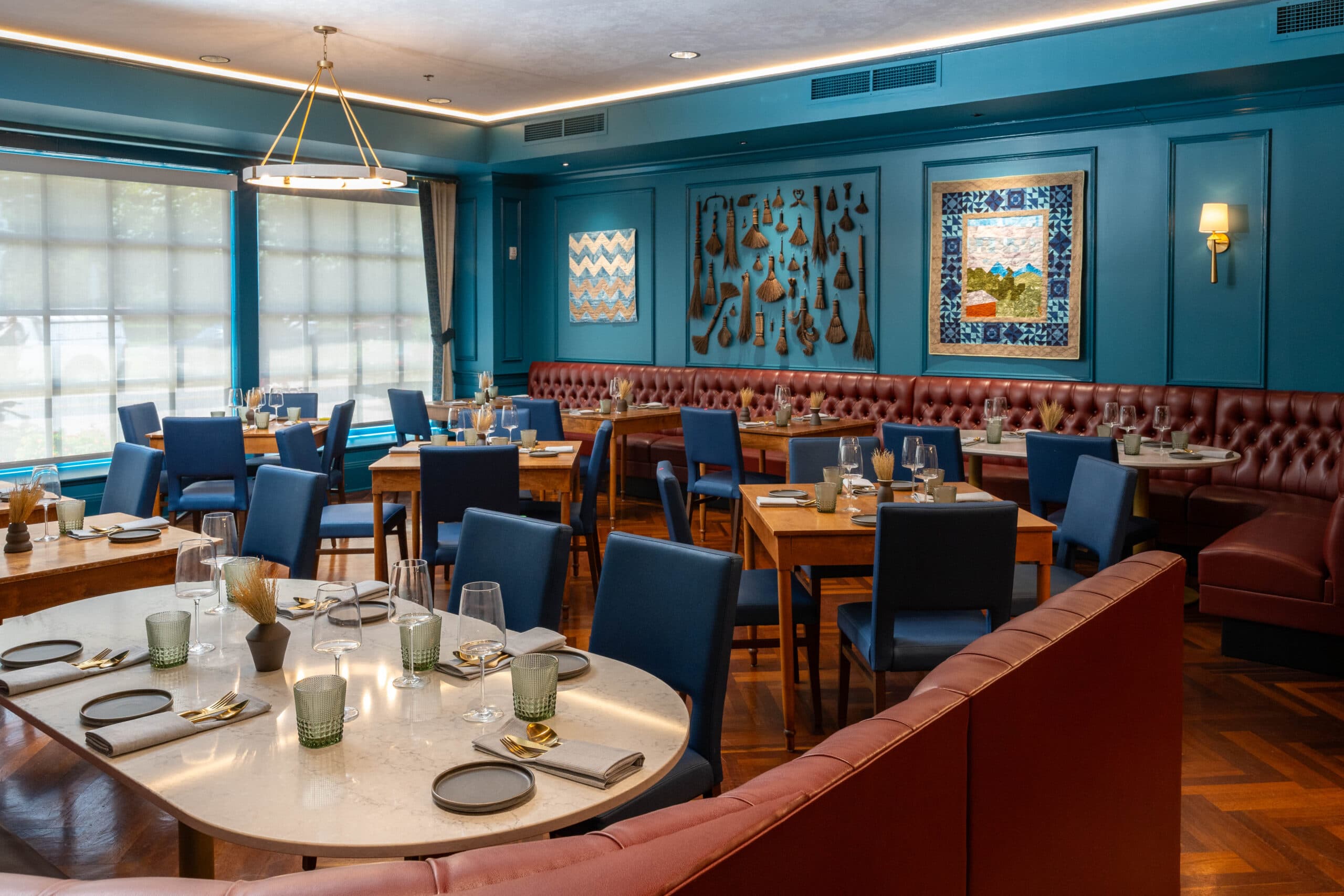 A room with blue walls, deep red booths, dining chairs, and tables neatly laid with cutlery is brightly shown in warm lighting. Large, hanging quilts and a broomcorn display, both crafted by students, are on display on the wall in the background.