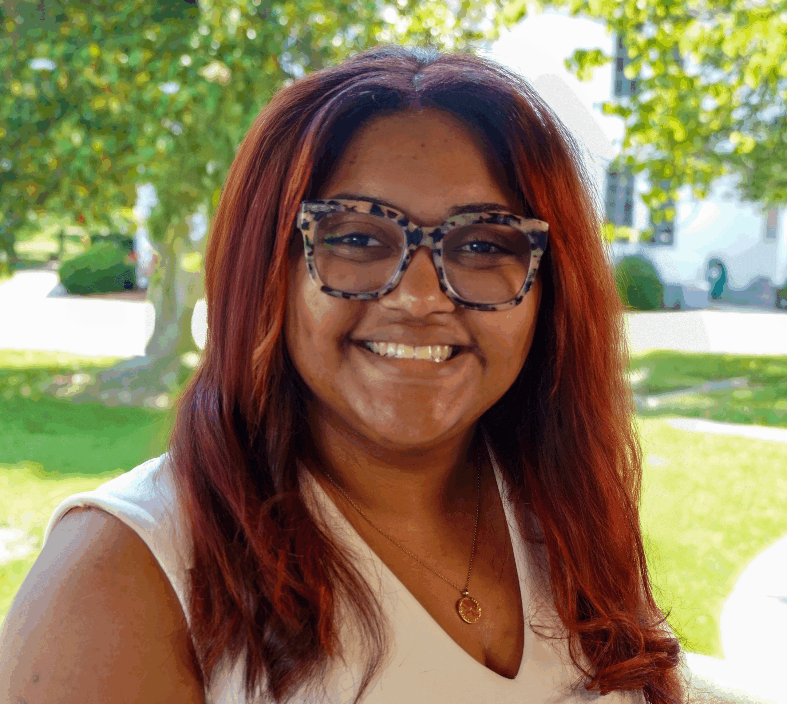 A photo of Regional Admissions Manager for the Ohio River Valley region, Jaleesa Nunez, standing outside amongst green grass and trees, wearing a white v-neck sleeveless sweater, smiling.