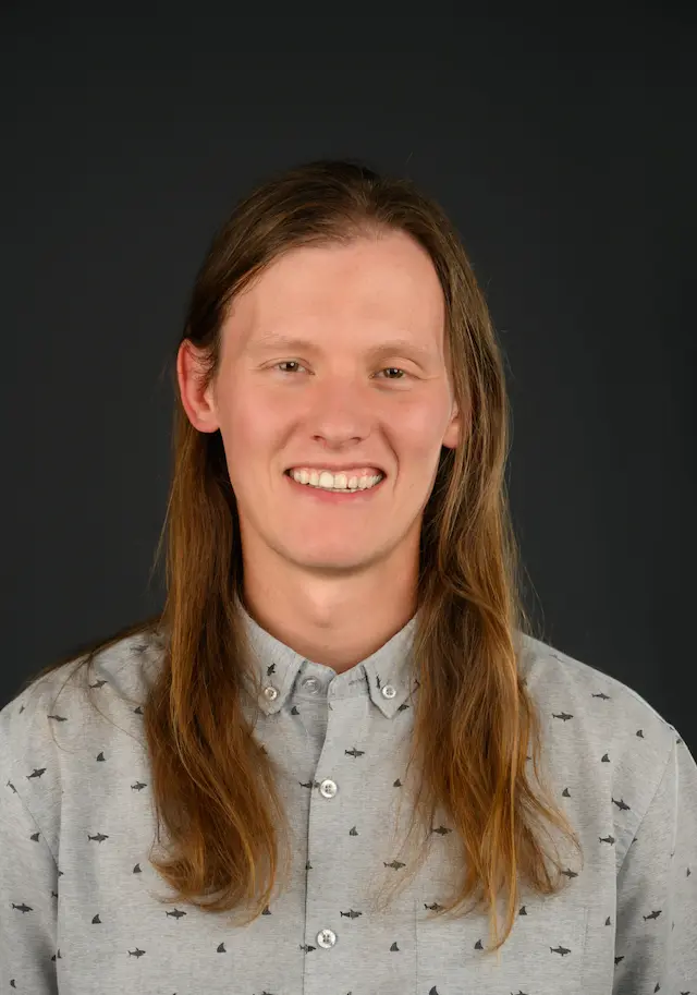 Keiffer Williams portrait wearing a patterned shirt with long hair smiling and a grey background.