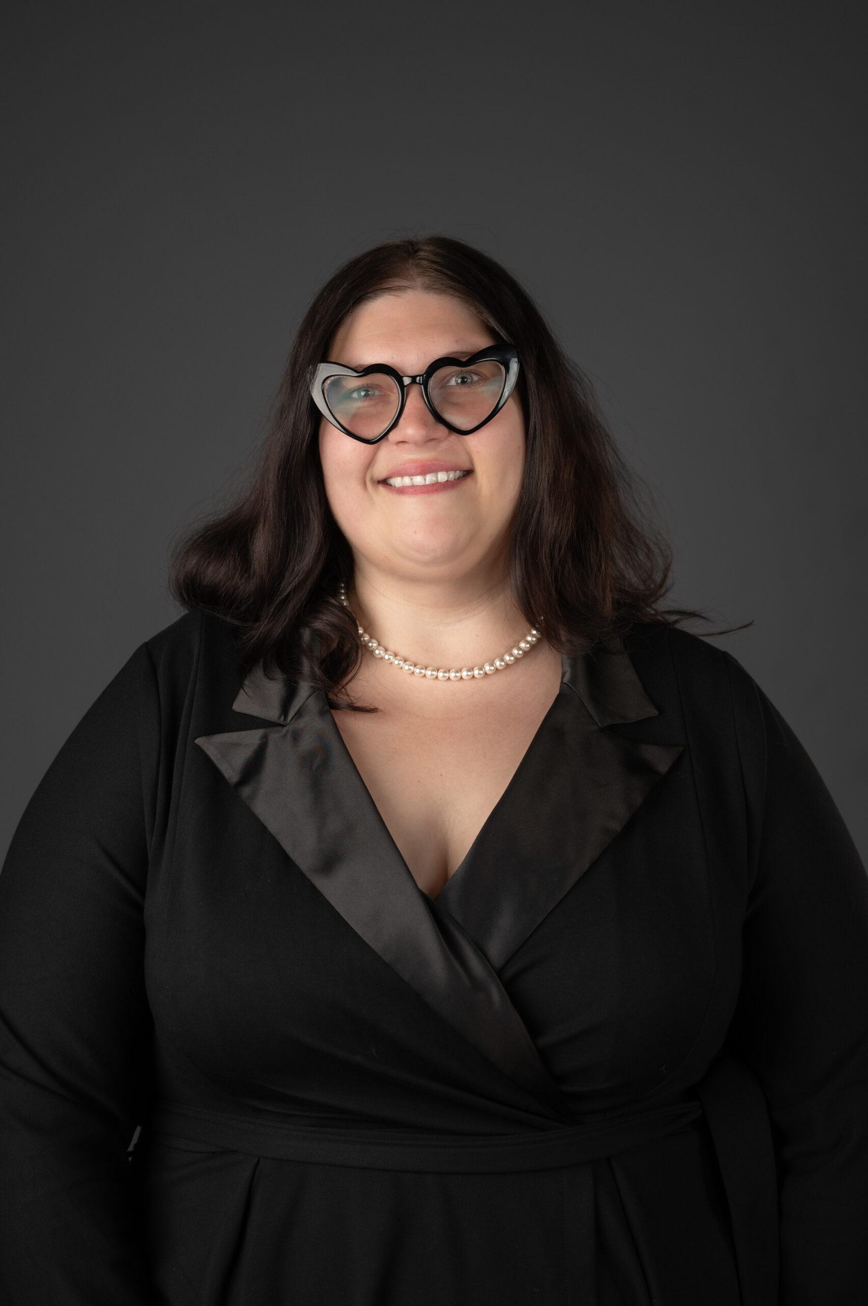 Sam Cole portrait wearing a black dress with black glasses smiling and behind is a grey background