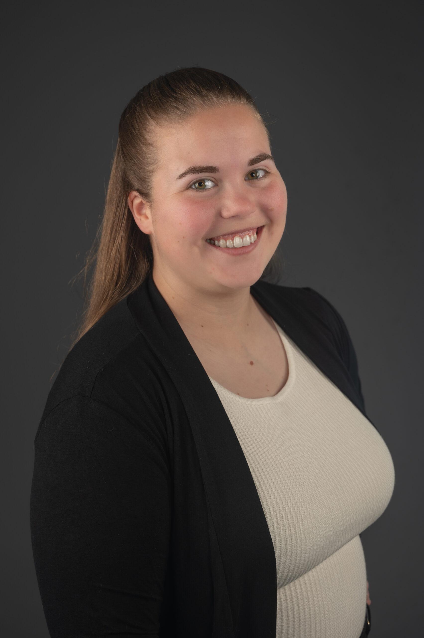 Jaedyn Rawson portrait smiling to the camera wearing a caramel top and a cardigan with grey background.