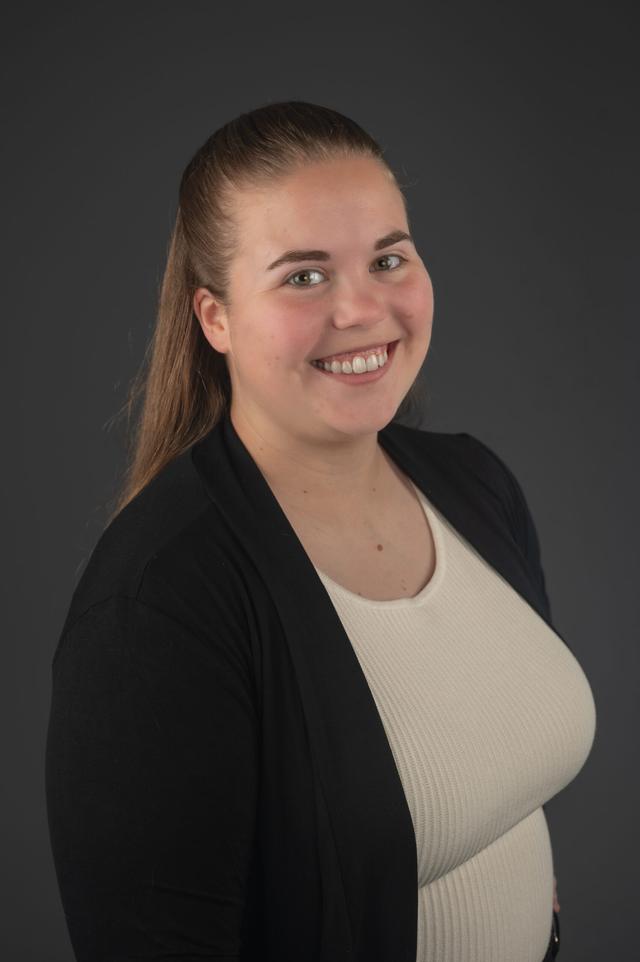 Jaedyn Rawson portrait smiling to the camera wearing a caramel top and a cardigan with grey background.