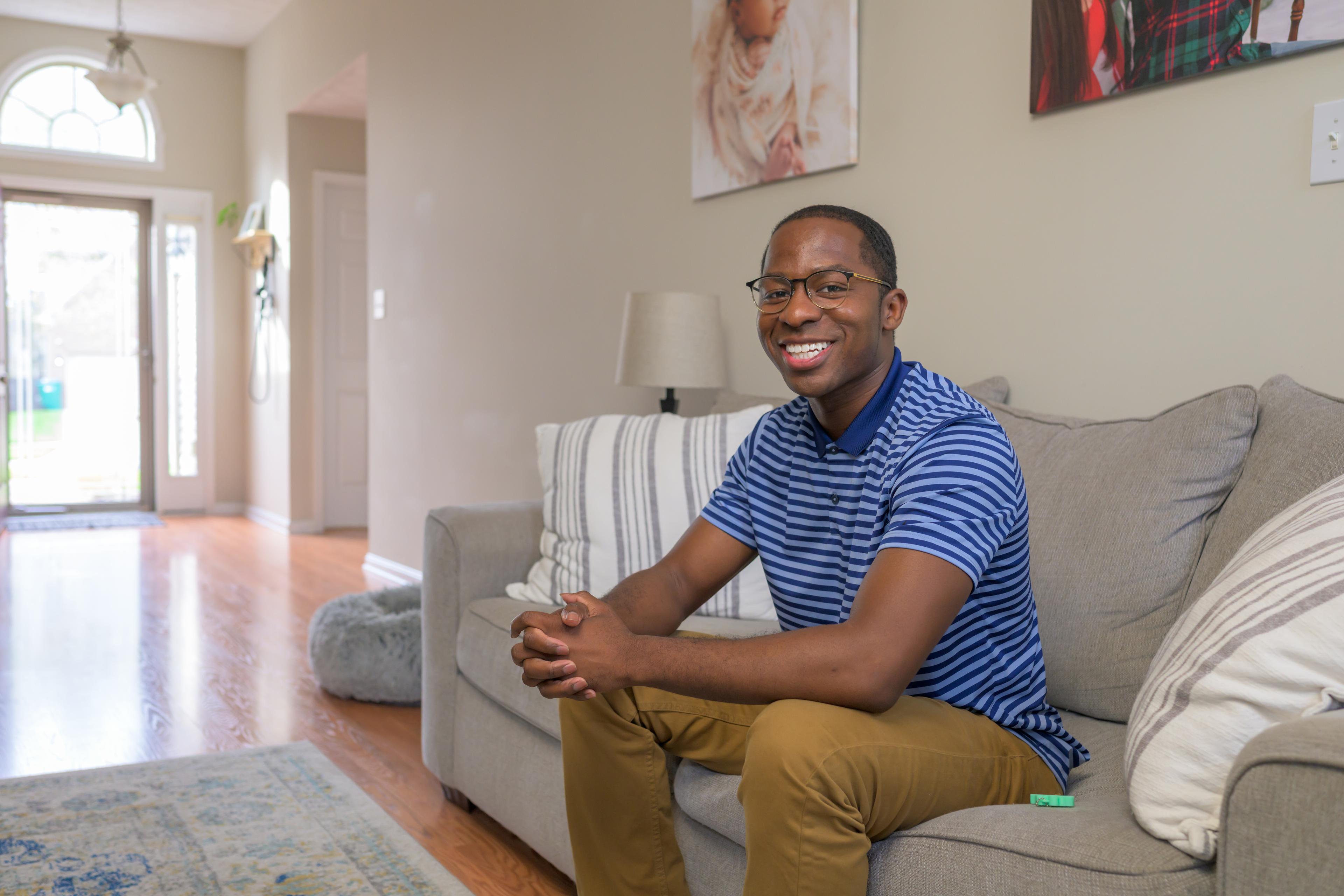 A smiling man wearing a striped blue shirt sits on a couch and looks to the camera.