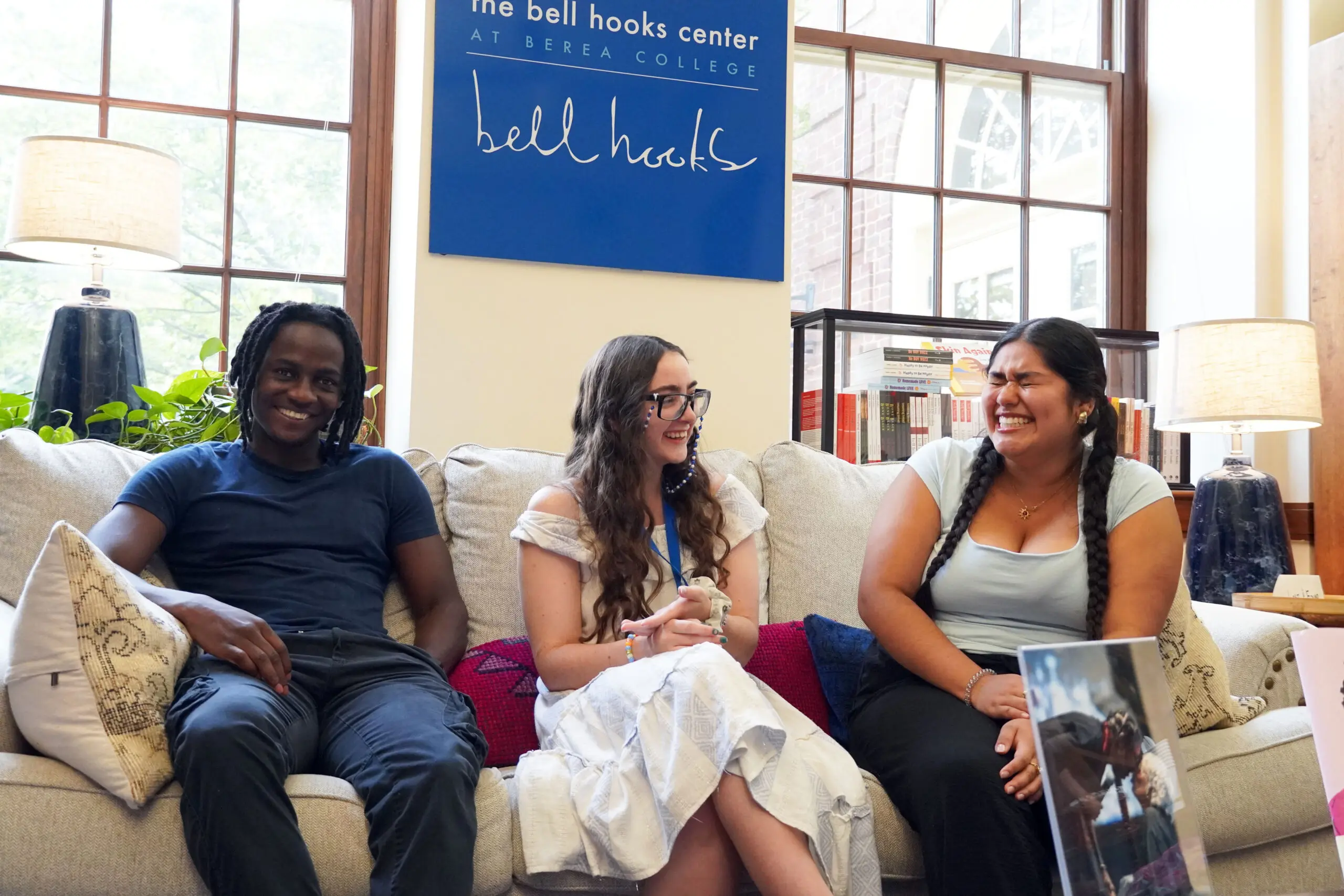 Three students sit together on a light-colored sofa in a bright lounge space at Berea College’s bell hooks center. Large windows, table lamps, plants, and bookshelves create a welcoming atmosphere. A blue wall sign above the sofa reads, “The bell hooks center at Berea College.” The individuals appear engaged in conversation, with an open laptop visible in the foreground.