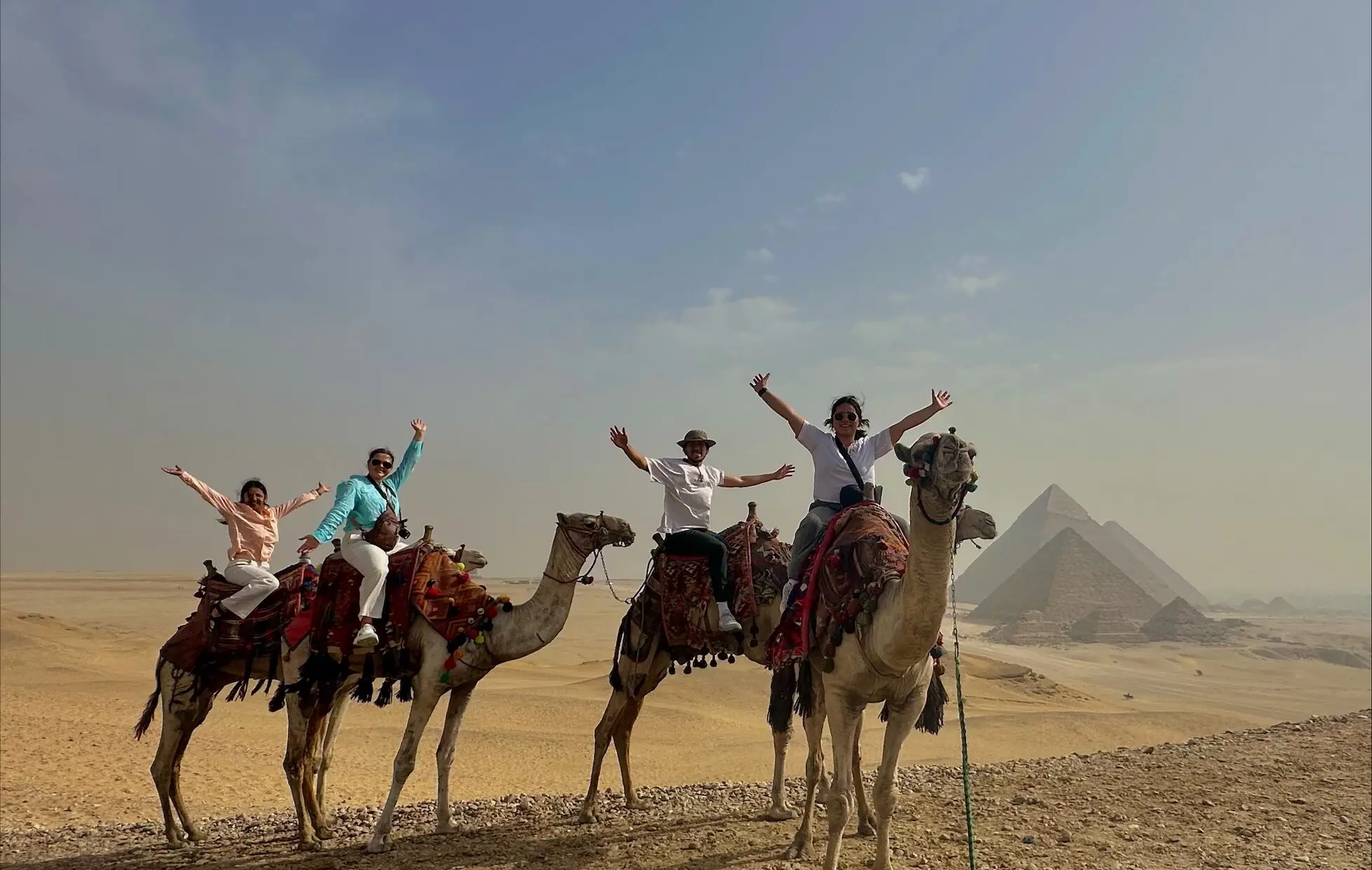Four people ride camels across a desert plateau near the Pyramids of Giza. Each rider raises their arms while seated on colorfully decorated saddles. The pyramids stand in the background under a hazy blue sky, with expansive sand and rocky terrain stretching across the landscape.