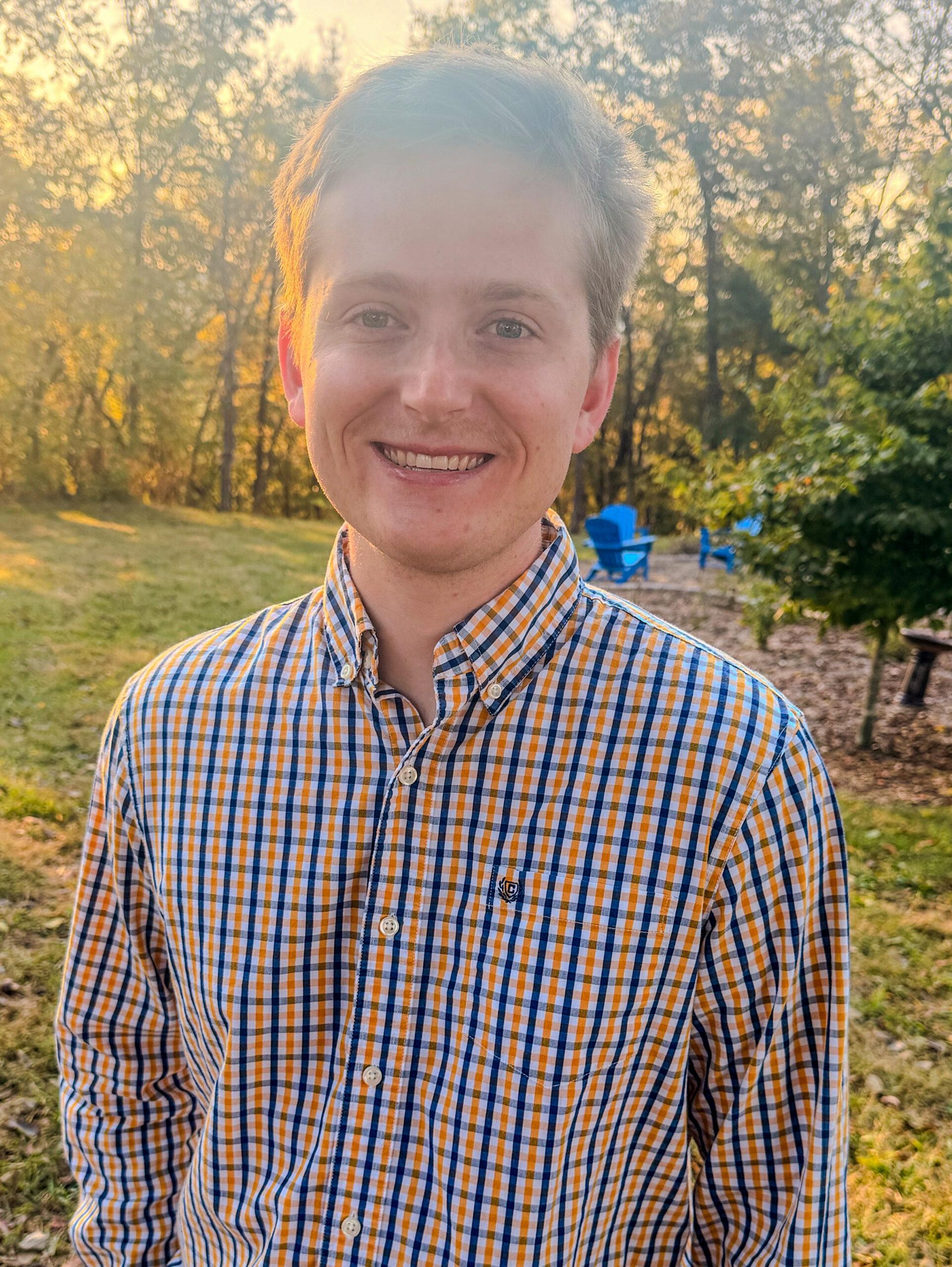 Dawson Rule wearing plaid button-down shirt standing outdoors in golden-hour light, with trees and grassy grounds in the background.